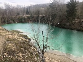 Paisaje natural con un lago turquesa cristalino, rodeado de árboles y terreno rocoso.