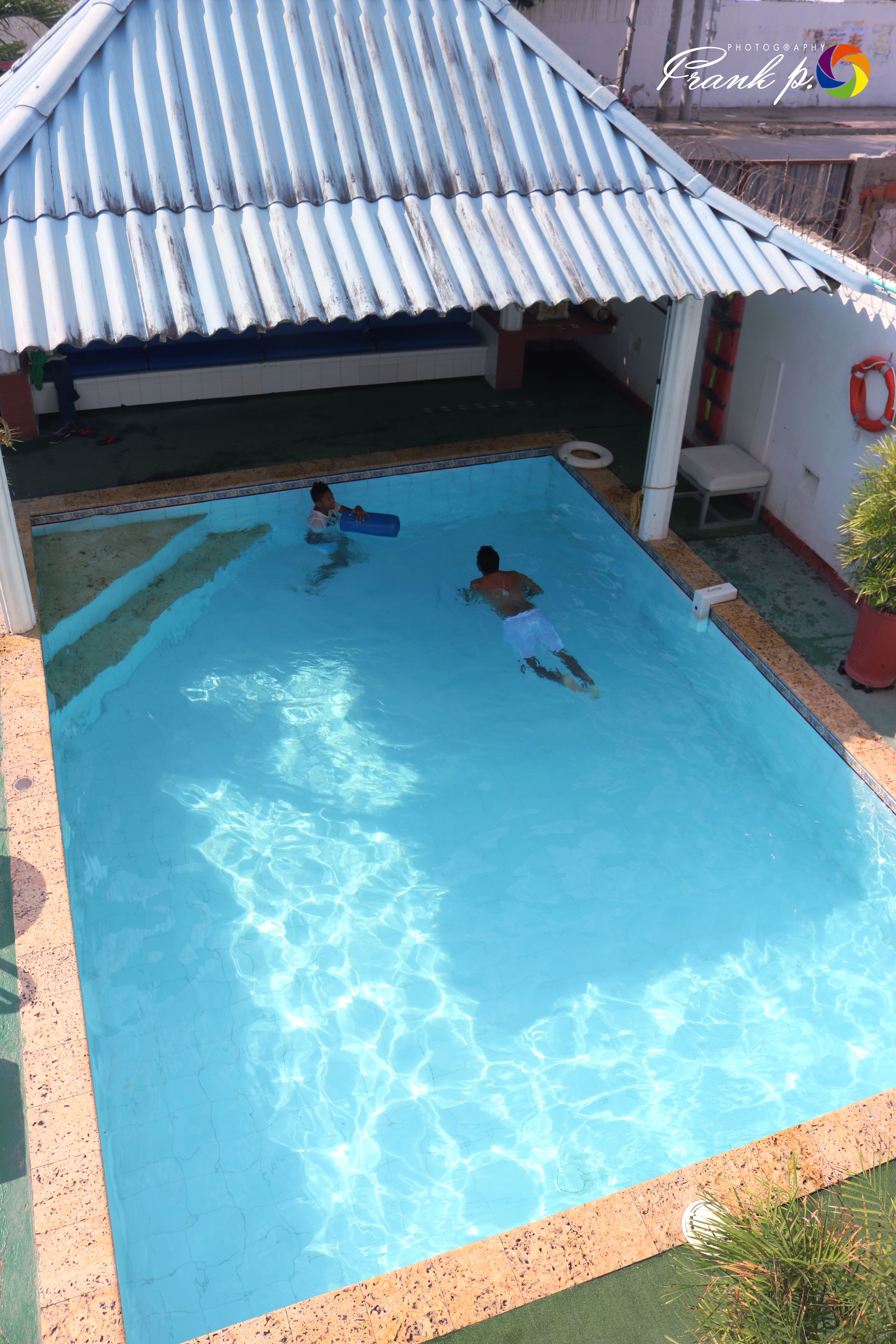 Une piscine où se trouvent deux personnes, une zone abritée et une bouée de sauvetage fixée au mur.
