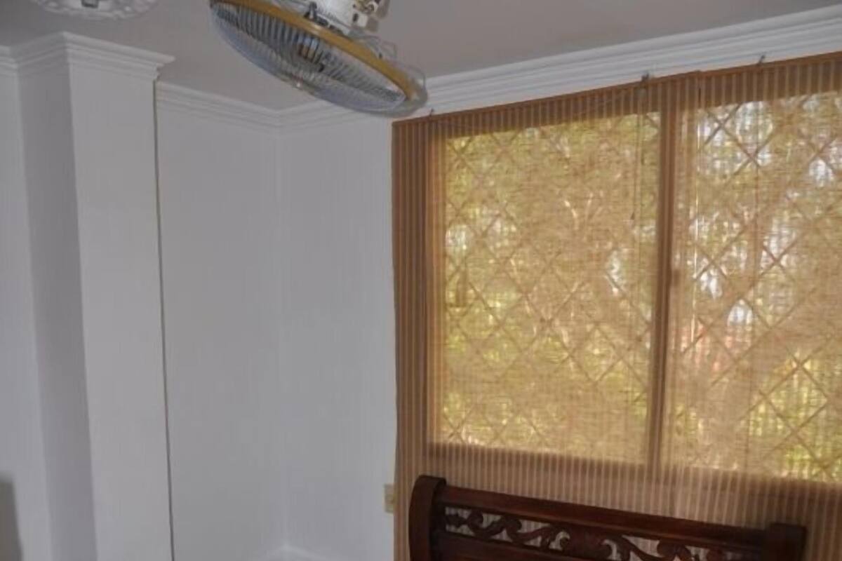 Une chambre à coucher avec un lit en bois, une table de chevet, un ventilateur de plafond et une fenêtre avec des stores.