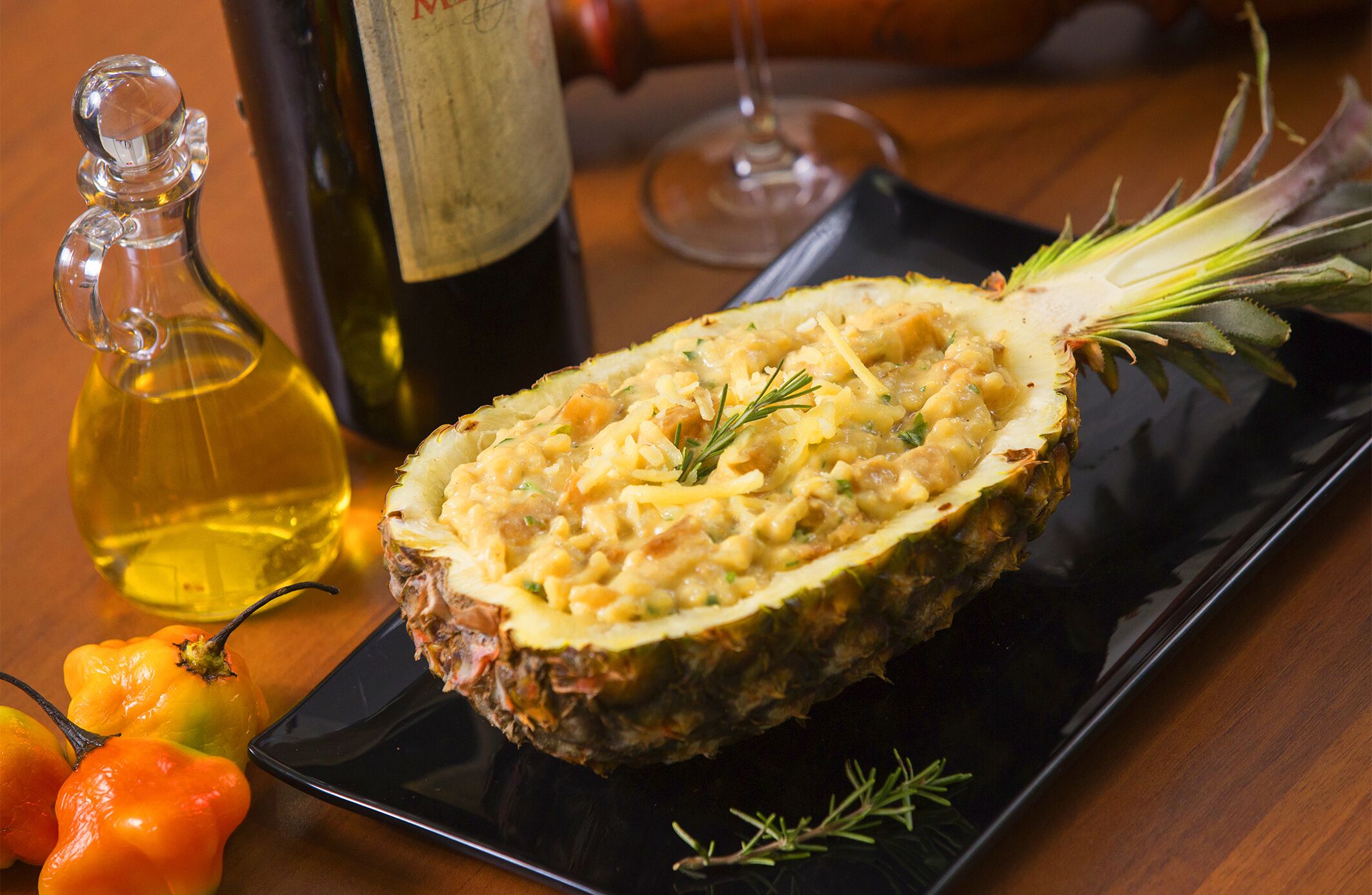A dish of stuffed pineapple on a black plate, with a bottle of olive oil and a wine glass in the background.