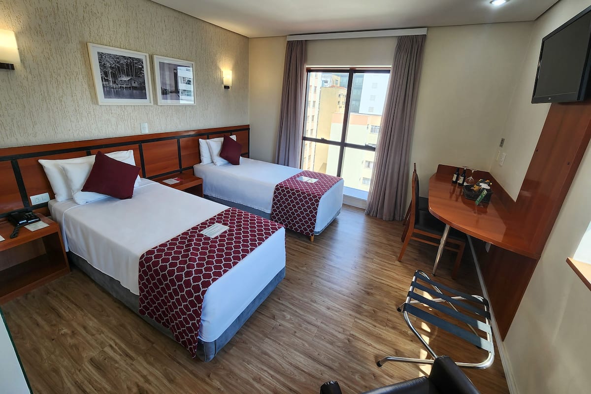 Ein Hotelzimmer mit zwei Betten, einem Schreibtisch, einem Stuhl, einem Fernseher und einem fenster mit Vorhängen.