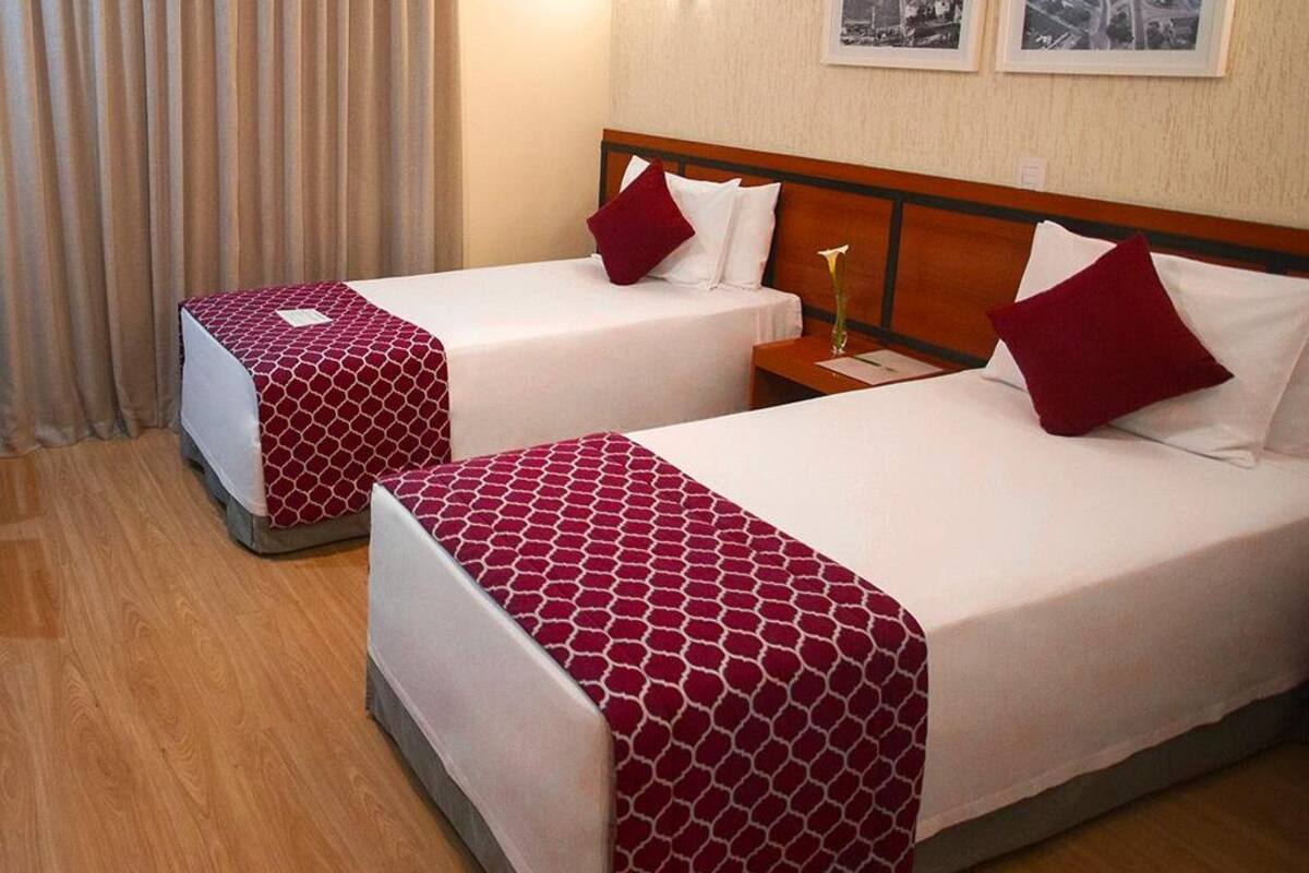 Ein Hotelzimmer mit zwei Einzelbetten, jeweils mit gemusterten Bettdecken und Kissen. Es gibt ein hölzernes Kopfstück, einen Nachttisch mit einer Lampe sowie ein Fenster mit Vorhängen.
