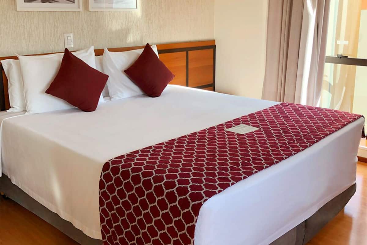 Ein Hotelzimmer mit einem großen Bett, weißer Bettwäsche und einer rot gemusterten Tagesdecke. Darüber an der Wand befinden sich zwei gerahmte Bilder.