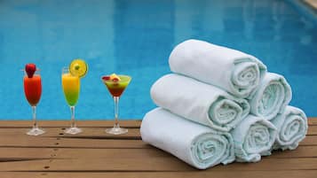 A wooden table with three colorful cocktails and a stack of neatly rolled white towels by a pool.