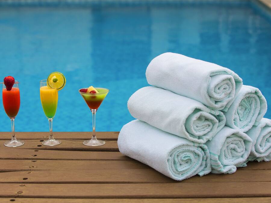 A wooden table with three colorful cocktails and a stack of neatly rolled white towels by a pool.