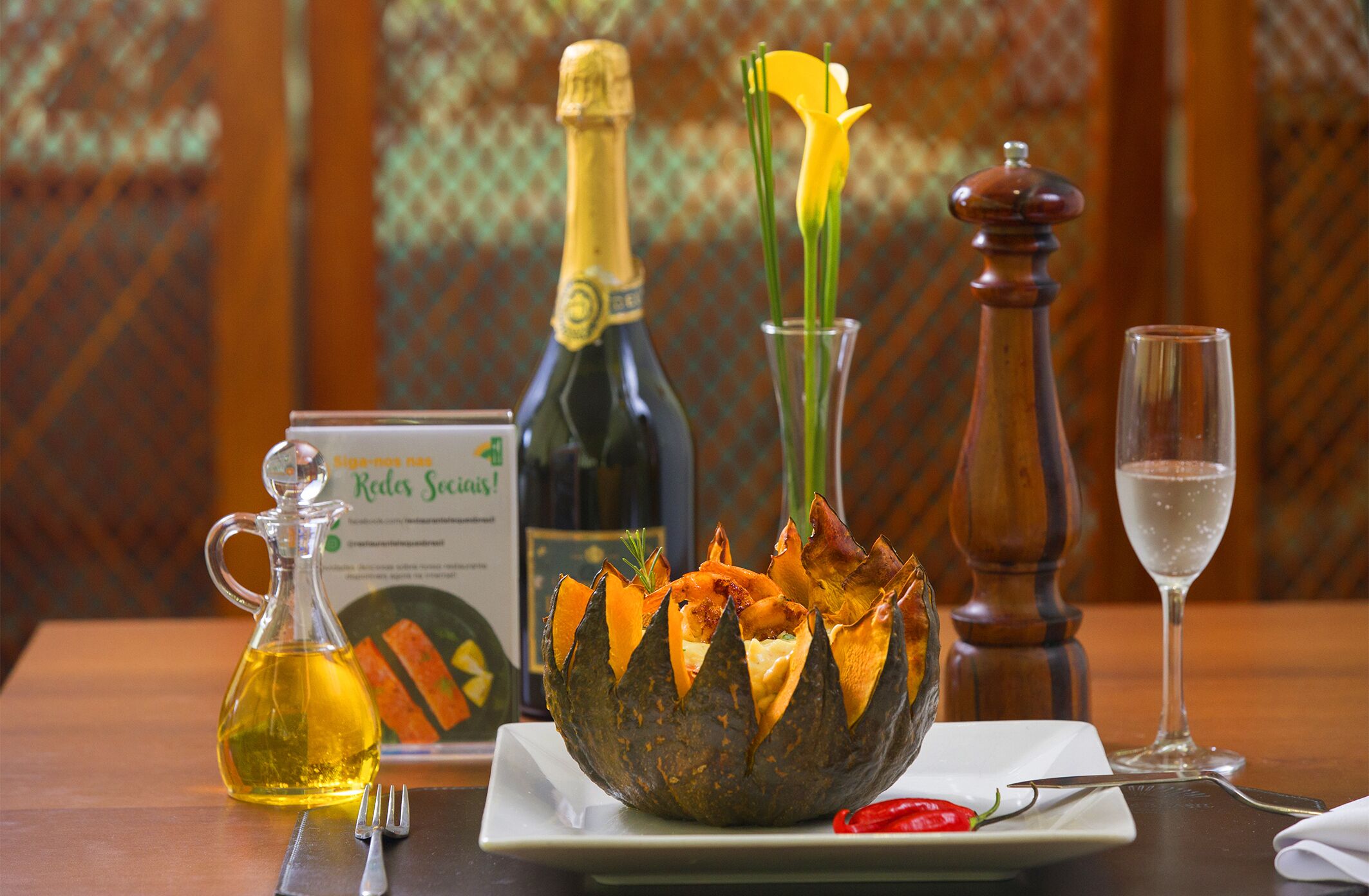 A table setting with a bottle of champagne, a glass of water, a salt and pepper shaker, a bottle of olive oil, a plate with a red pepper, and a decorative centerpiece.