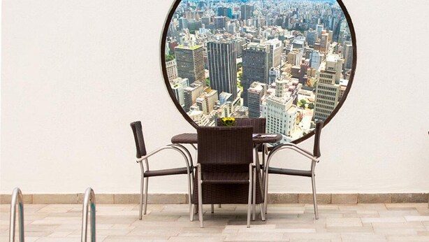 A poolside area with a view of a cityscape through a circular window, two wicker chairs, and a small table with a vase.