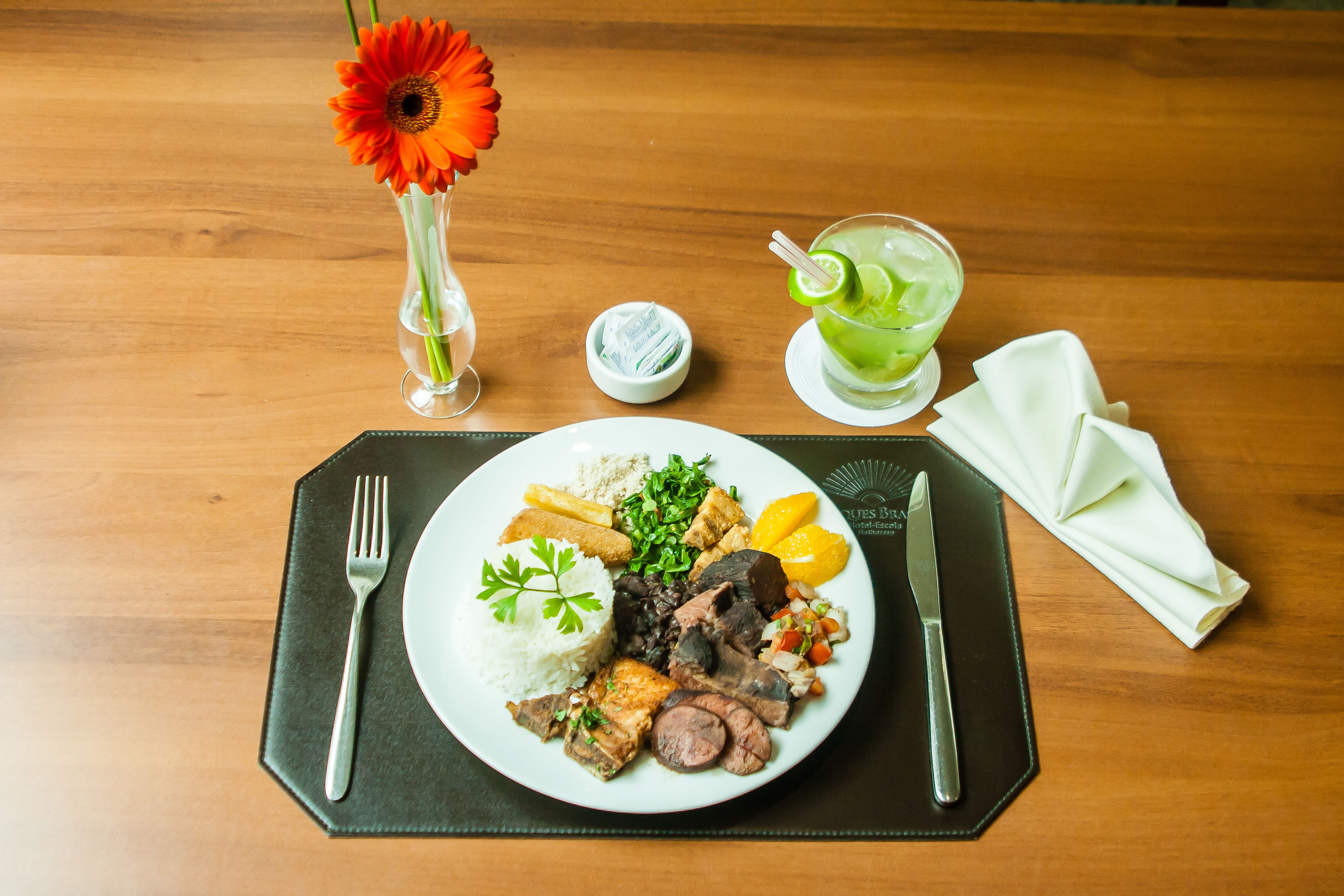 A plate of food with rice, meat, and vegetables, a glass of green drink, a flower in a vase, and a napkin on a wooden table.