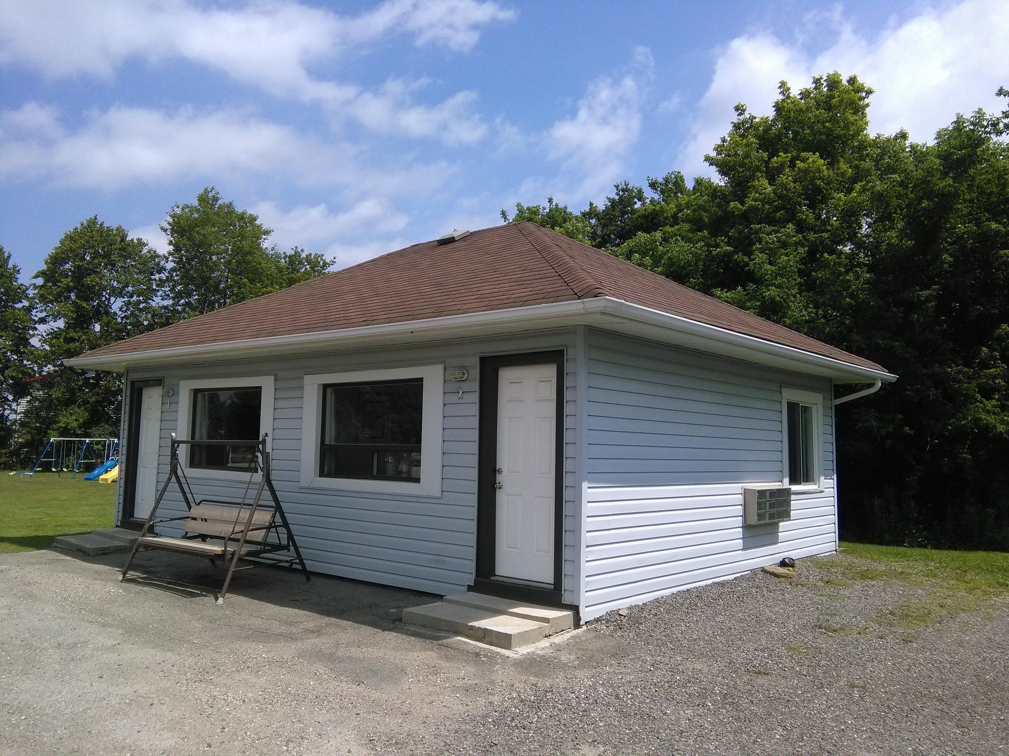 A small, single-story building with a brown roof and white siding, featuring a front door, a window, and a small porch area.