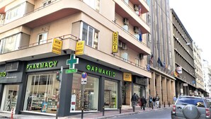 A pharmacy with a green cross sign, a sign for 'AMBROSIA SUITES & APARTMENTS', and multiple flags on the building. - Ambrosia Suites & Aparts (Athens)