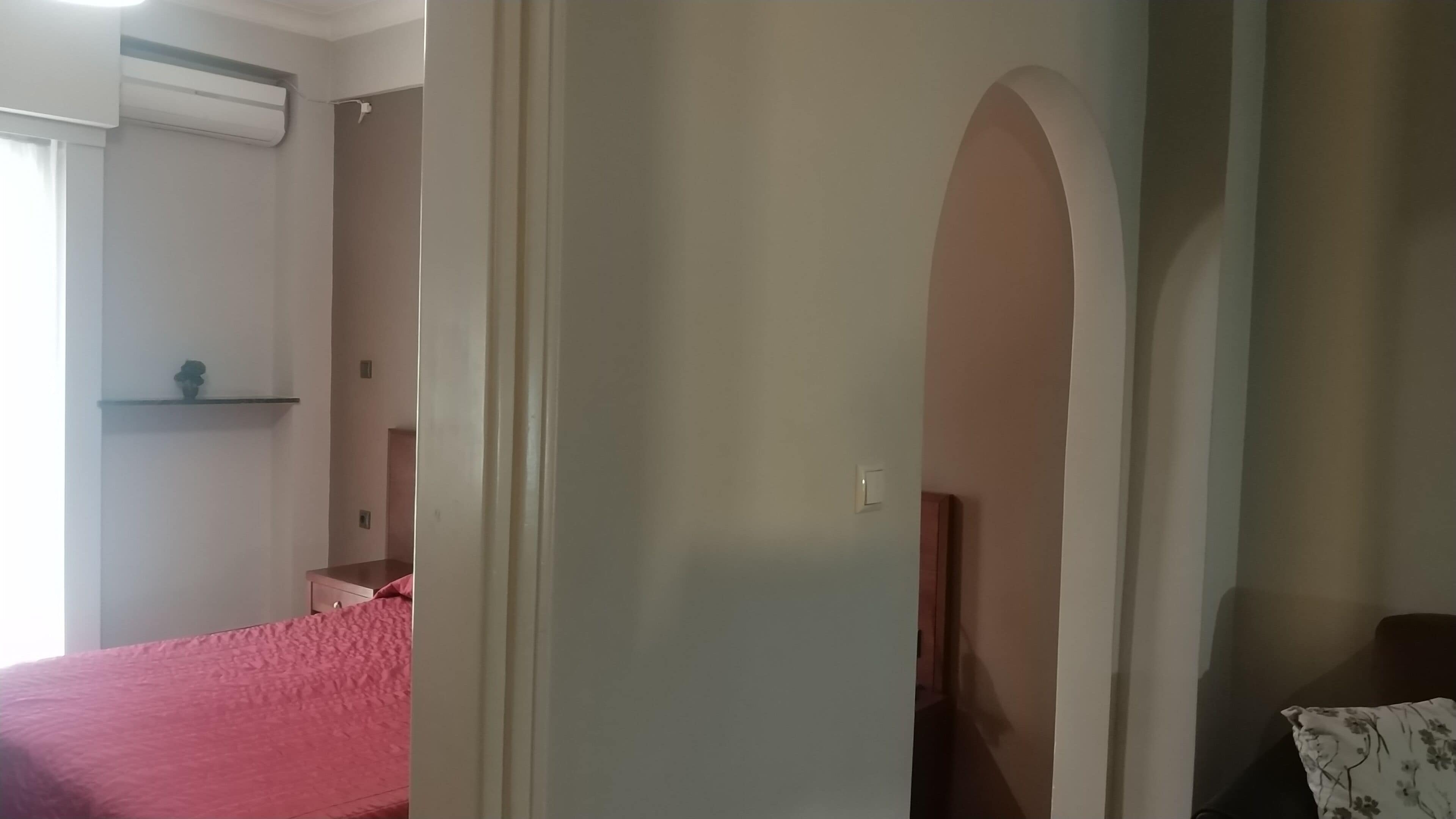 A bedroom with a bed, a nightstand, and a dresser. There is a window with curtains and an air conditioning unit on the wall.