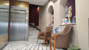 An elevator, two brown leather chairs, a marble-patterned floor, and a wall with a statue and travel brochures. - Ambrosia Suites & Aparts (Athens)
