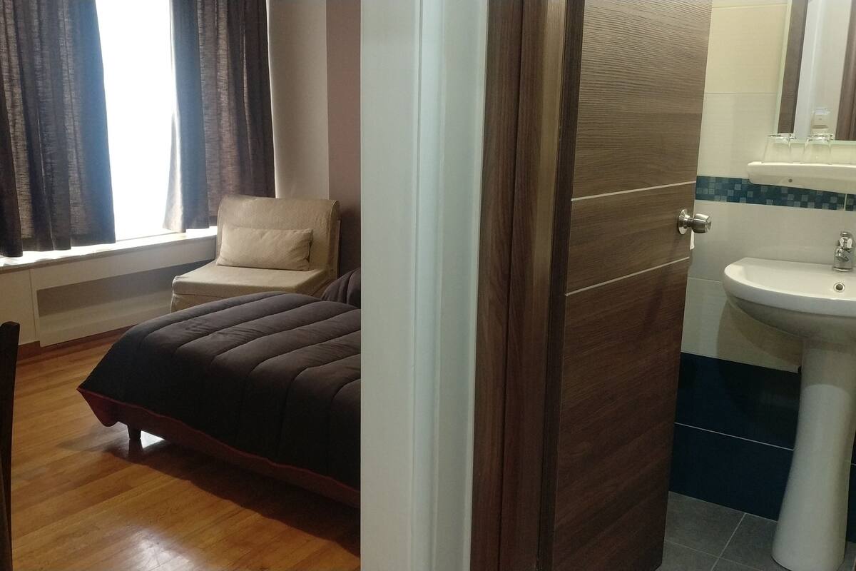 A bedroom with a bed, a chair, a window with curtains, and a bathroom with a sink and mirror.