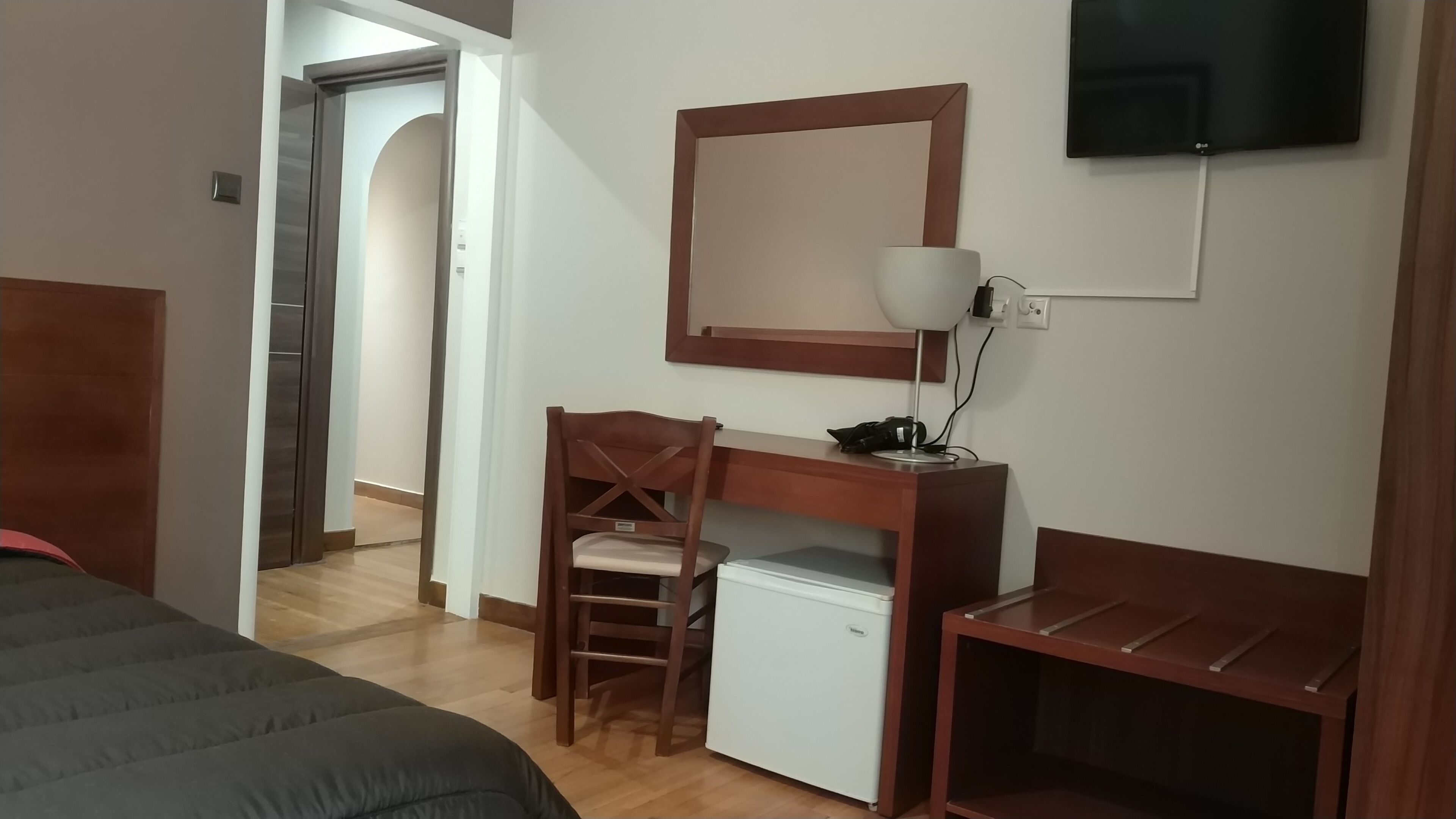 Ett hotellrum med en säng, ett skrivbord med en stol, ett litet kylskåp, en tv som är monterad på väggen och en träbänk.