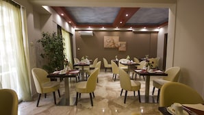 A dining area with multiple tables and chairs, a ceiling with recessed lighting, and a large plant. - Ambrosia Suites & Aparts (Athens)