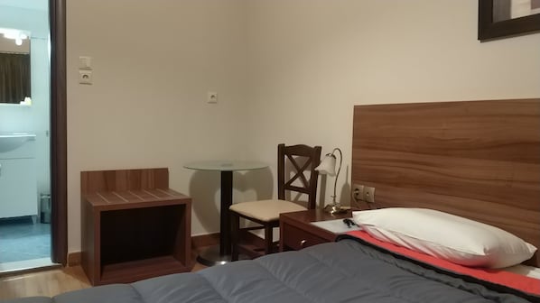 A hotel room with a bed, a wooden nightstand, a small table, a chair, and a lamp.