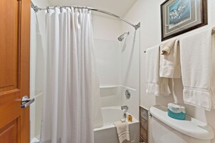Un baño con cortina de ducha, bañera, inodoro con toallero y un cuadro en la pared.