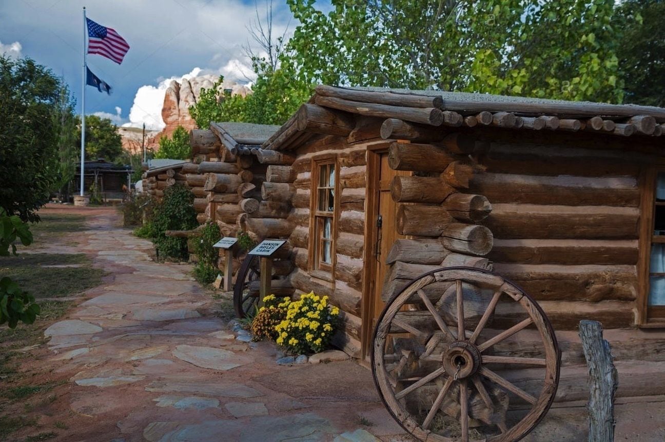 A rustic log cabin with a stone pathway leading to it, surrounded by greenery and a flag.