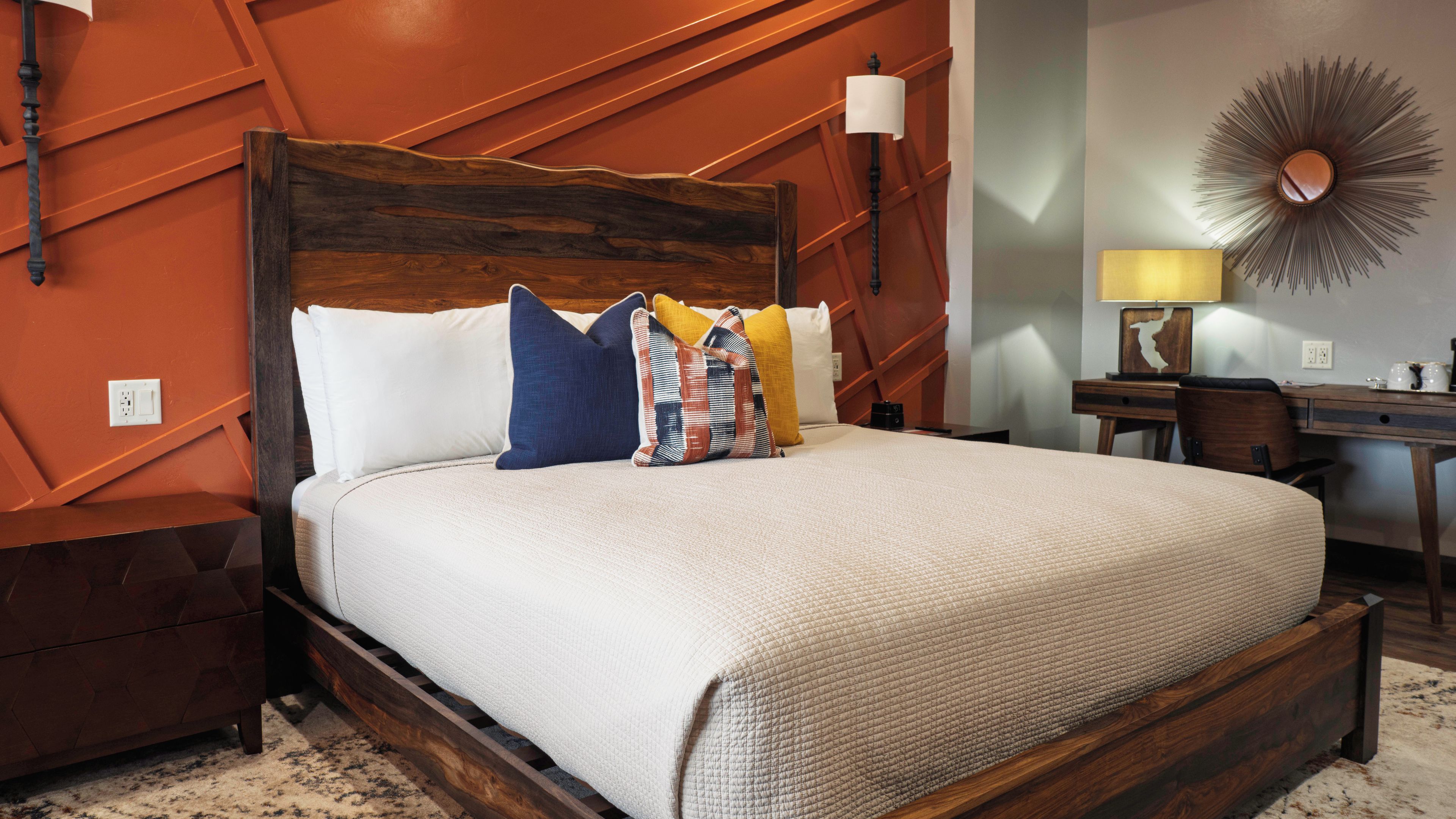 A bed with a wooden headboard, multiple pillows, a bedside table with a lamp, and a wall-mounted mirror.