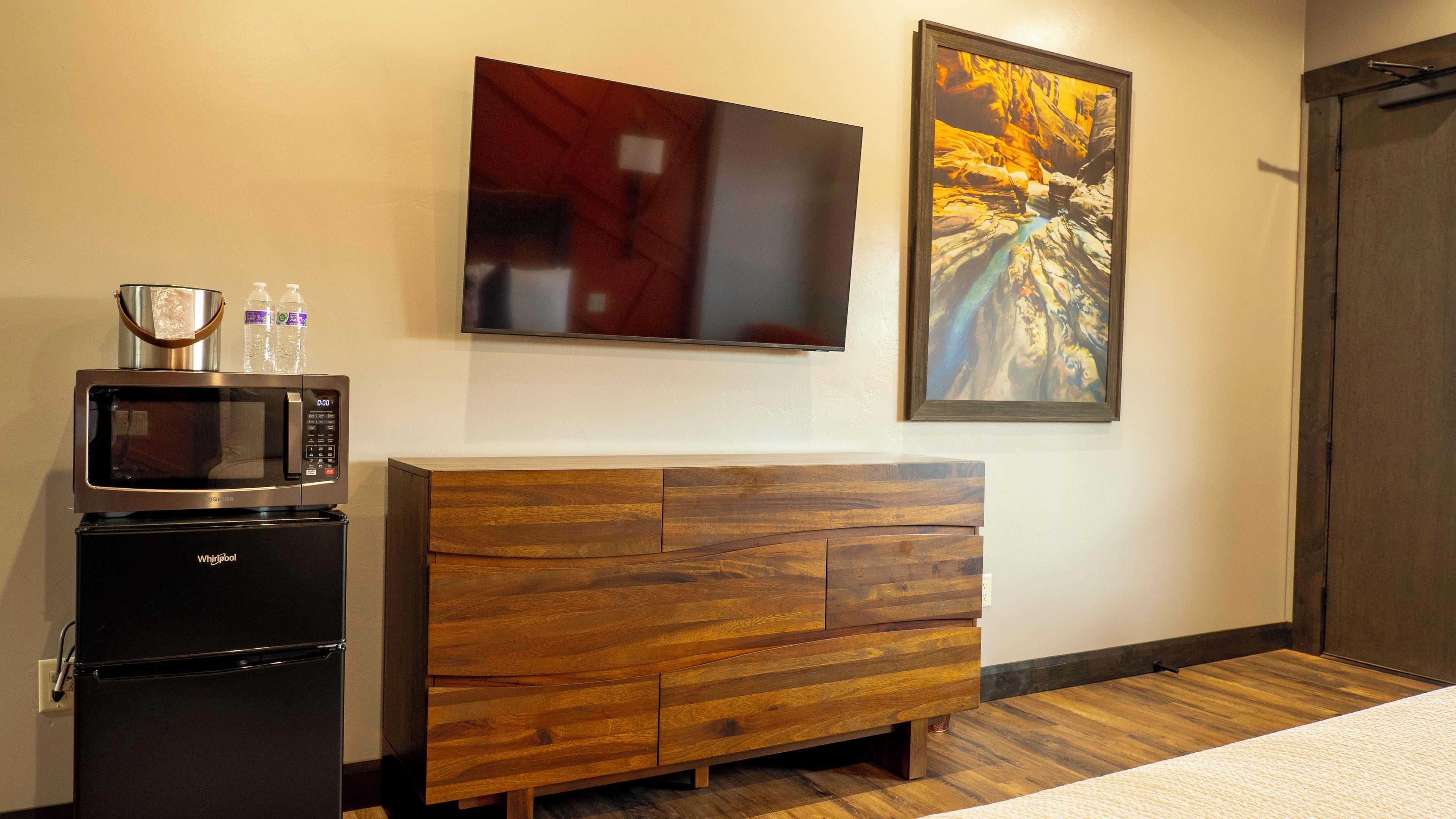 A hotel room with a microwave, refrigerator, wooden dresser, a flat-screen TV, and two framed artworks on the wall.