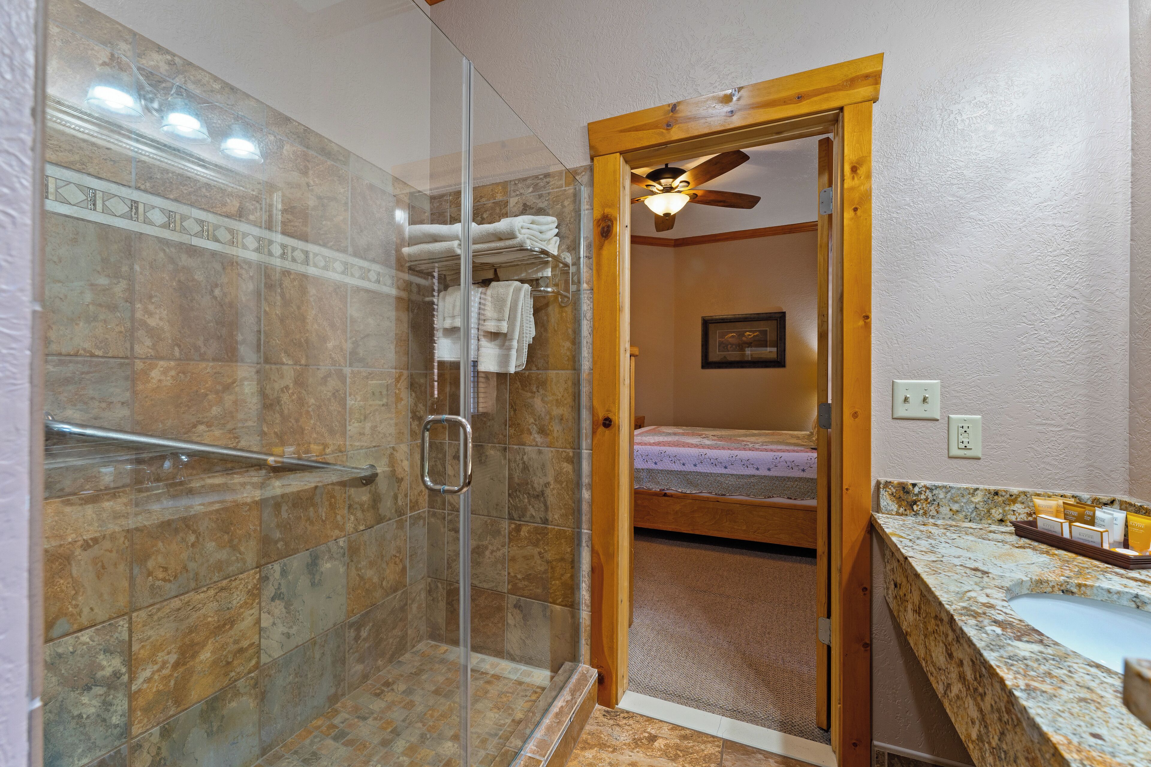 A bathroom with a glass shower enclosure, a wooden door leading to a bedroom, and a granite countertop with a sink.