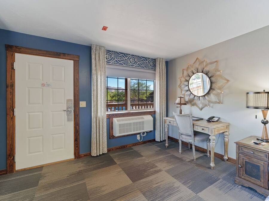 Une pièce avec un sol à carreaux, une porte blanche, un bureau avec une chaise, une table de chevet avec une lampe et un miroir décoratif accroché au mur.