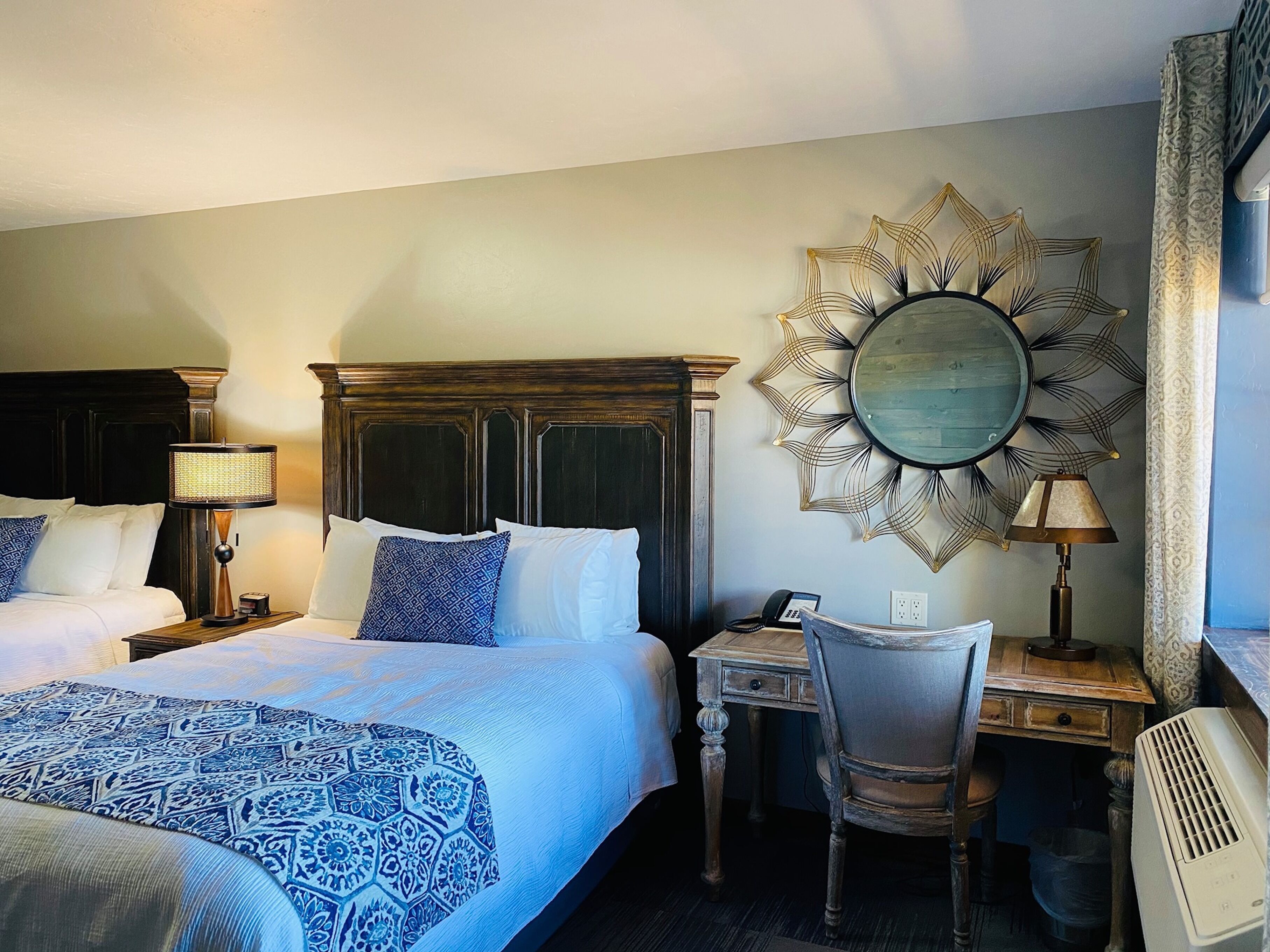 A hotel room with a large bed, a desk, a chair, a desk lamp, a telephone, and a decorative mirror.