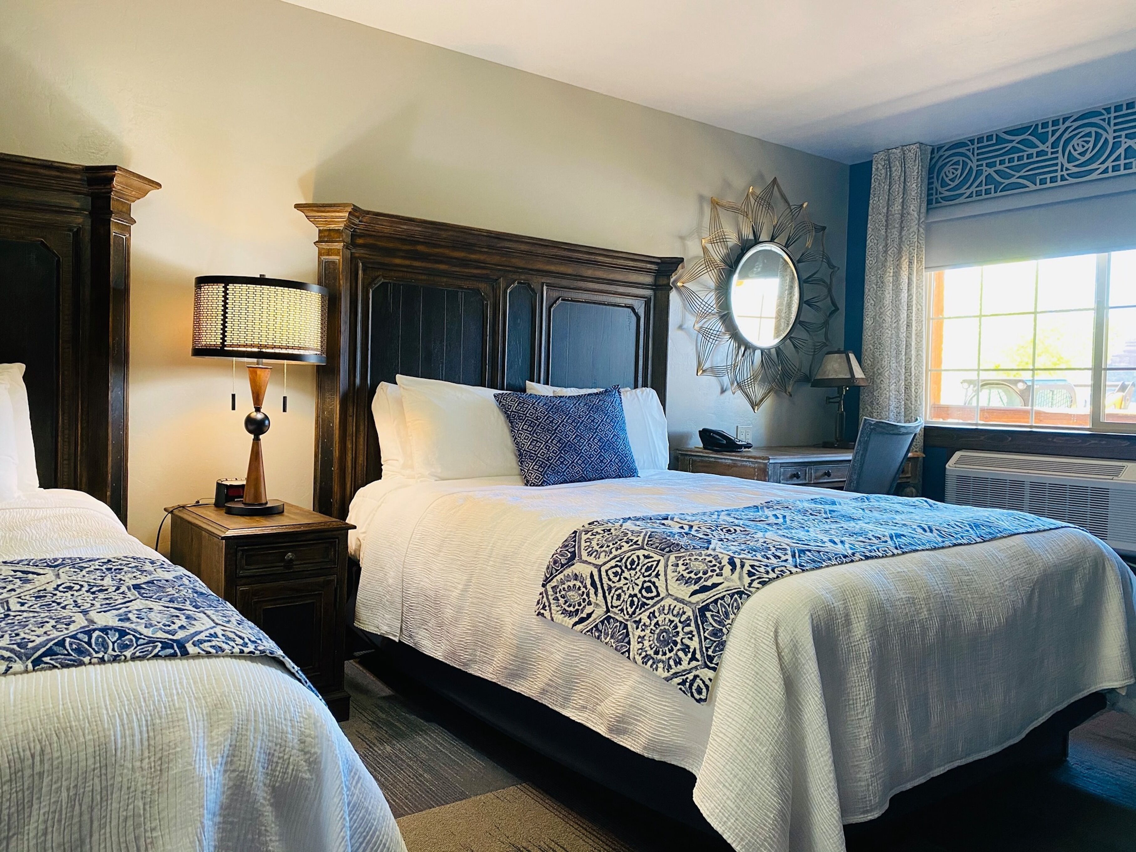 A hotel room with two beds, a large mirror, a decorative wall piece, and a window with curtains.
