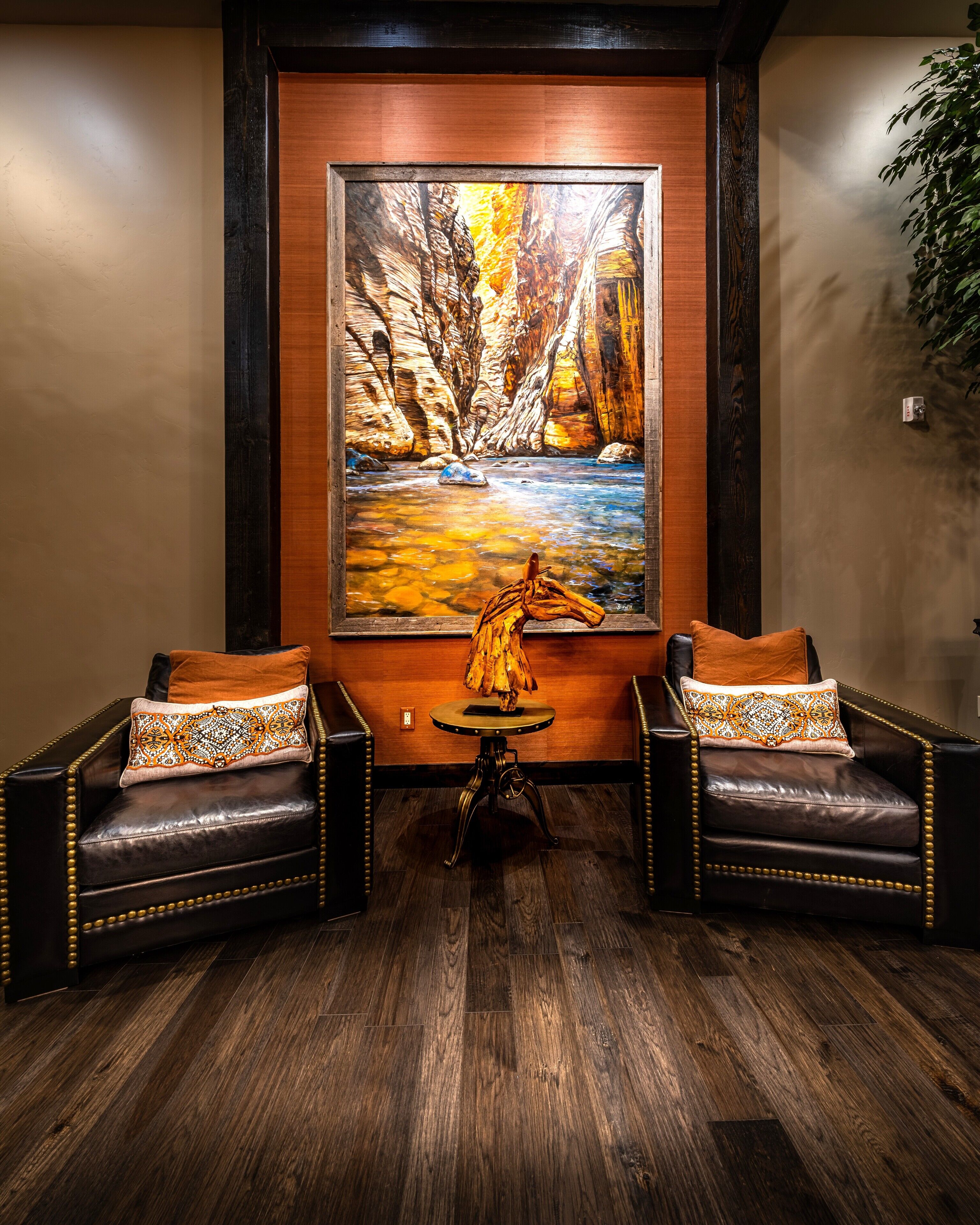 A room with a wooden floor, two leather armchairs with patterned cushions, a small table with a decorative horse sculpture, and a large painting of a canyon landscape on the wall.