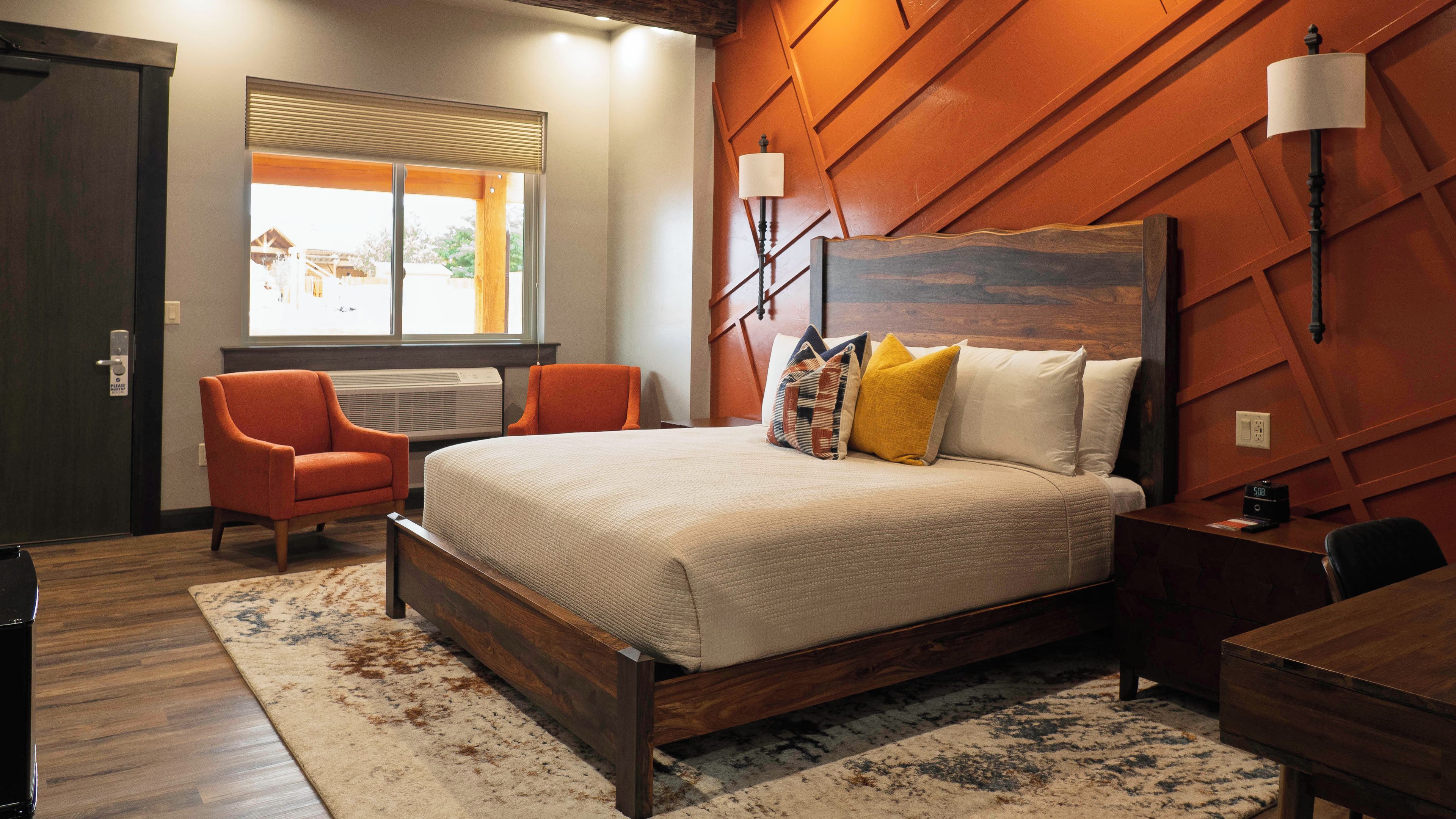 Une chambre à coucher avec une tête de lit en bois, un lit avec plusieurs oreillers, un bureau, une chaise et une fenêtre avec des stores.