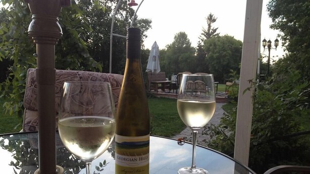 Une table en verre, avec deux verres à vin et une bouteille de vin, disposée à l’extérieur, avec vue sur un jardin.