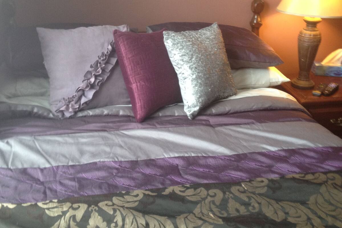 Een net opgemaakt bed met een bedsprei met dessin, een lijst met een abstract schilderij erboven, en een bedlampje.