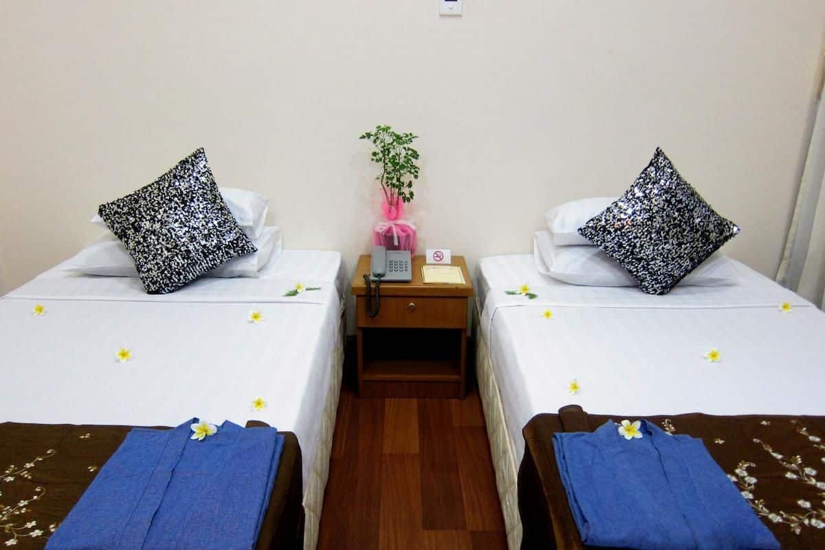 Quarto de hotel com duas camas de solteiro, cada uma com uma almofada e uma toalha azul. Há uma pequena mesa com um telefone e uma planta no centro.
