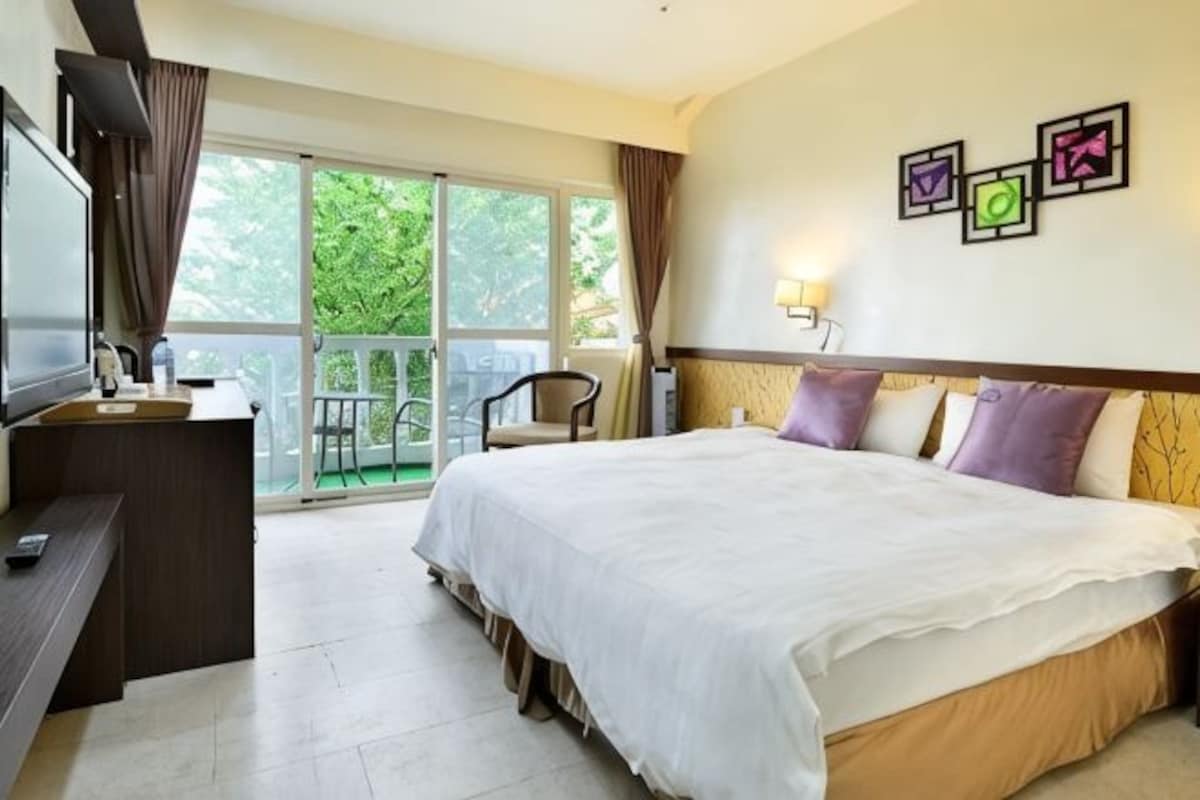 A hotel room with a bed, a desk, a chair, a TV, and a balcony with curtains.
