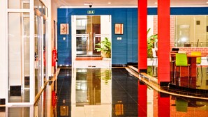 Un vestíbulo moderno con suelo negro reflectante, pared azul y pilares rojos. Hay un mostrador de recepción con sillas coloridas y una planta en maceta.