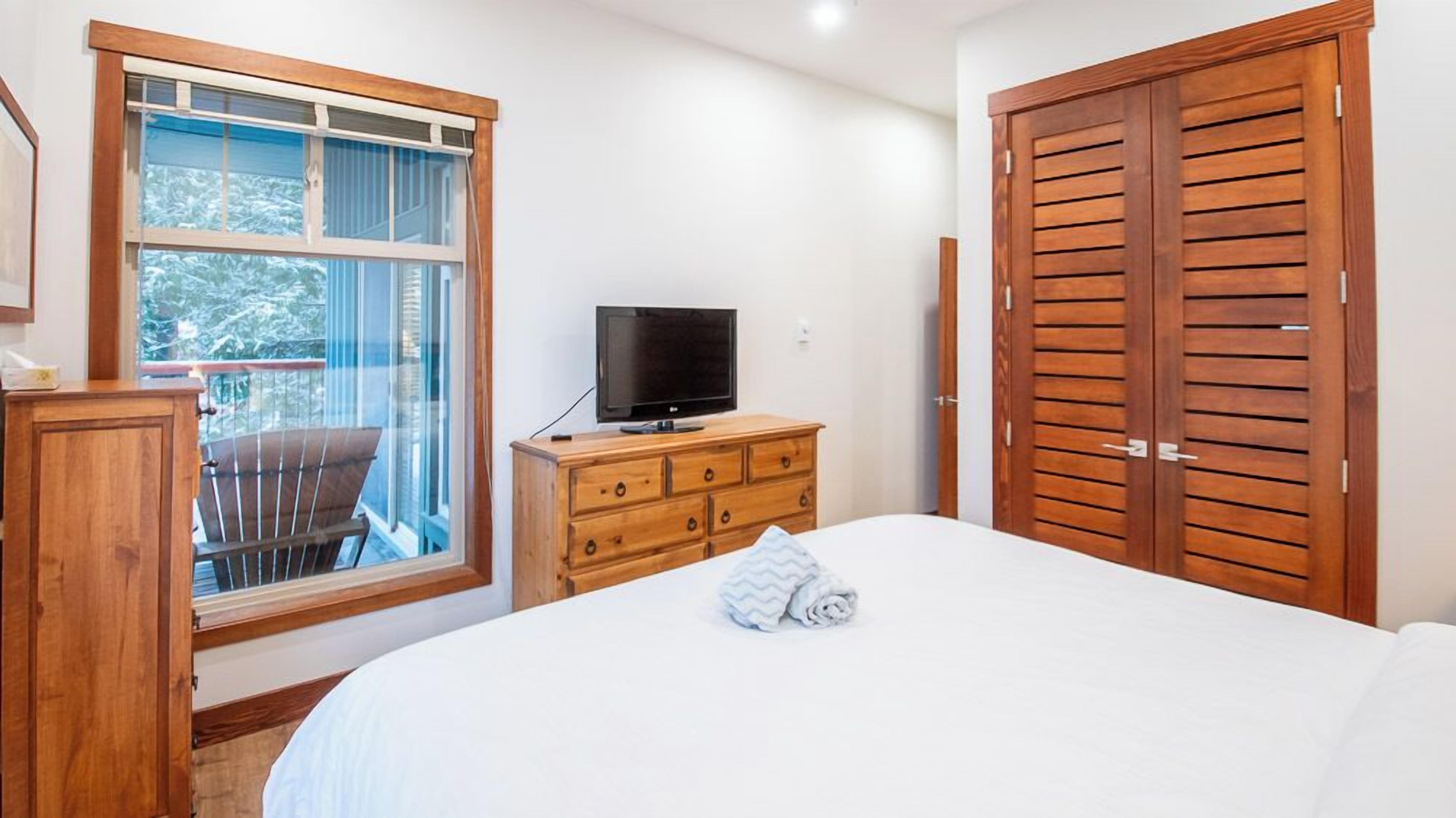Una camera da letto con un letto, una cassettiera in legno, una televisione e un'ampia finestra vista alberi.