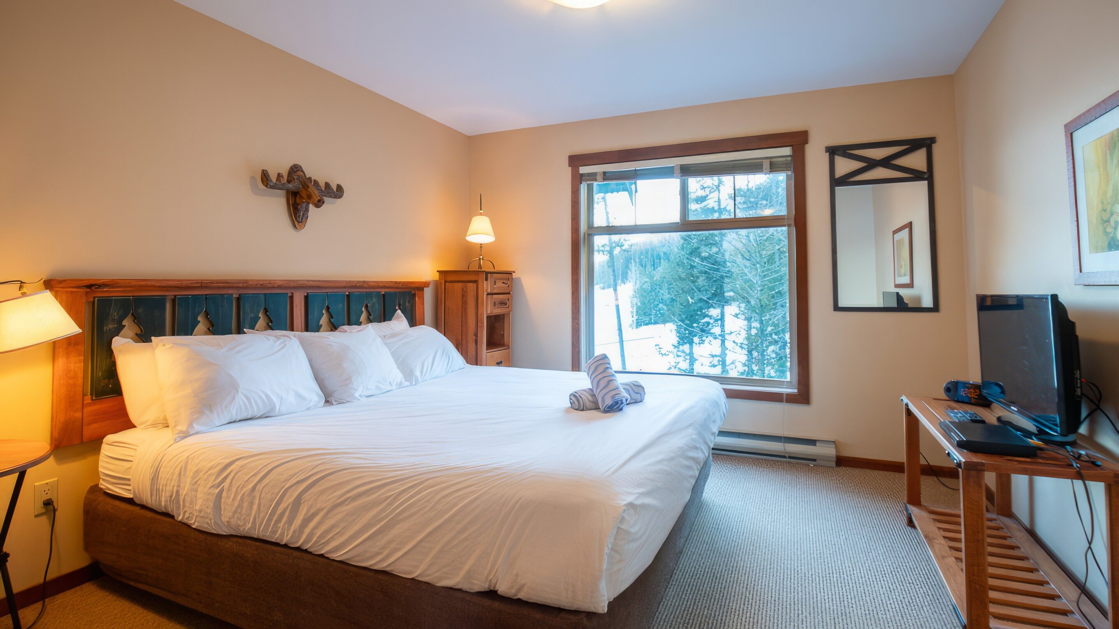 A bedroom with a large bed, a wooden headboard, a mounted animal head, a window with a view of trees, a desk with a computer, and a lamp.