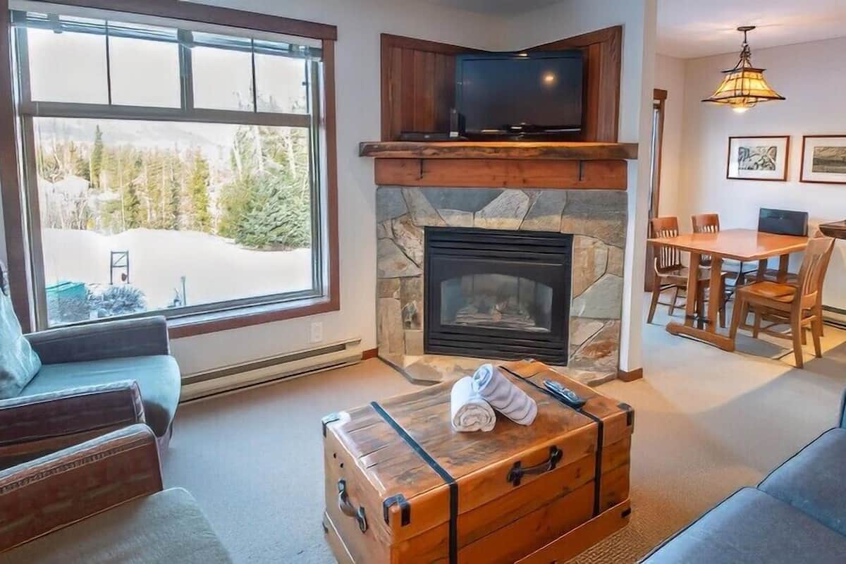 Ein gemütliches Wohnzimmer mit Kamin aus Stein, einem darüber angebrachten Fernseher und einem großen Fenster mit Blick auf eine verschneite Landschaft.