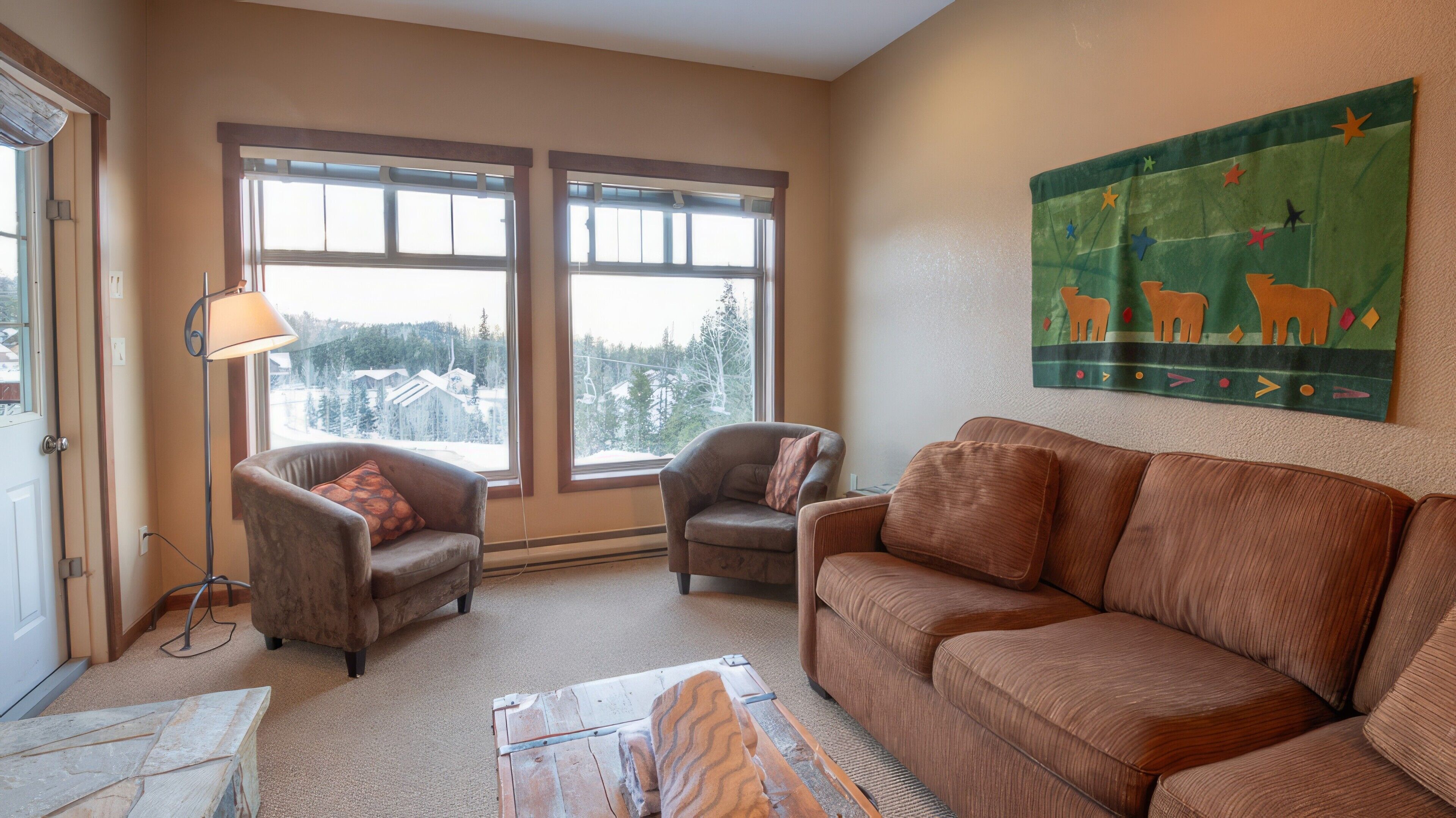 A cozy living room with a brown sofa, two armchairs, a floor lamp, and a painting of animals on the wall.