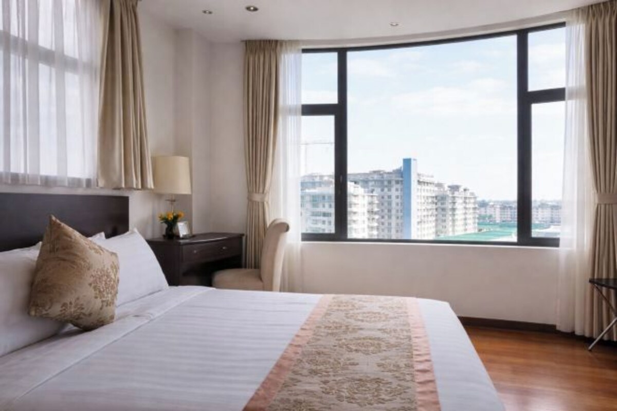 A hotel room with a large bed, a nightstand, a lamp, a chair, and a view of the city through a large window.