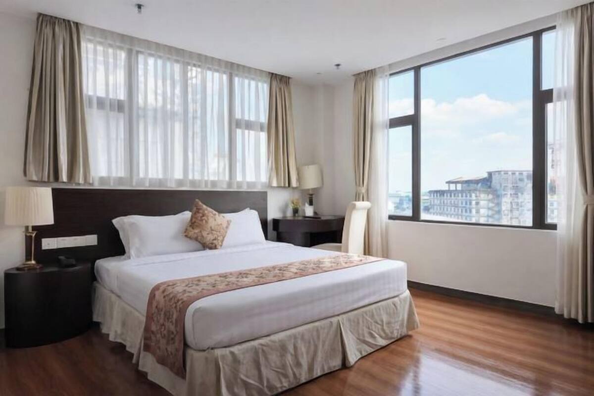 A hotel room with a large bed, a nightstand, a lamp, and a view of the city through large windows.