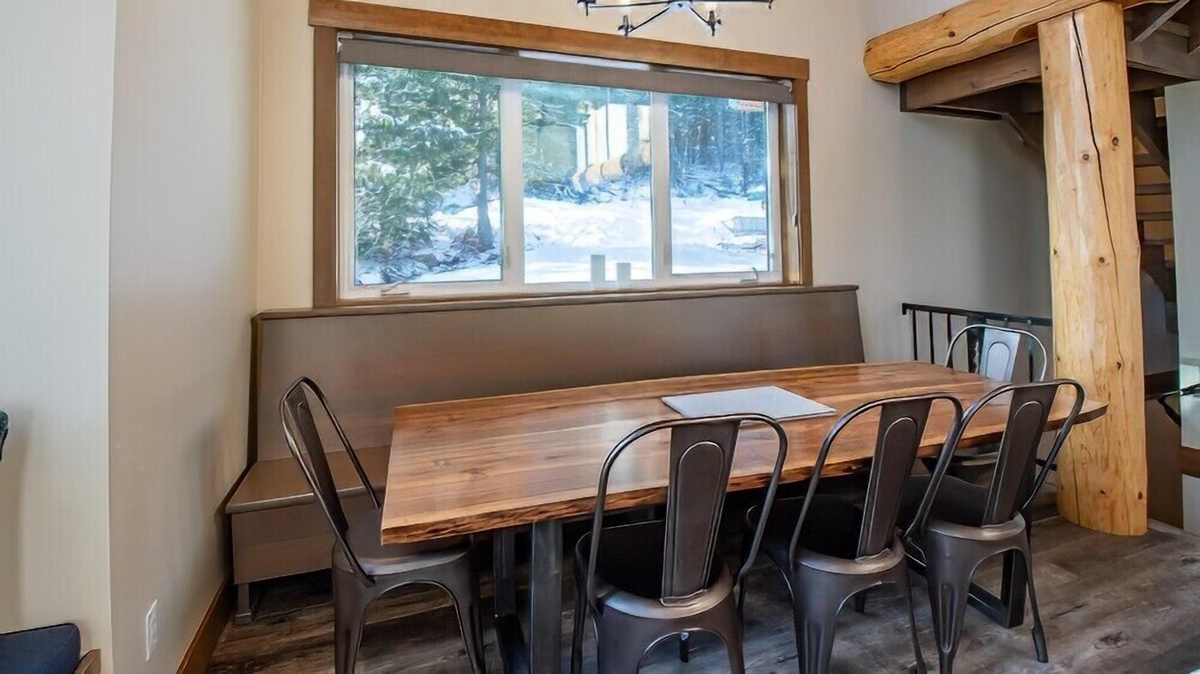 A dining area with a wooden table and chairs, a window with a view of trees, and a wooden staircase.
