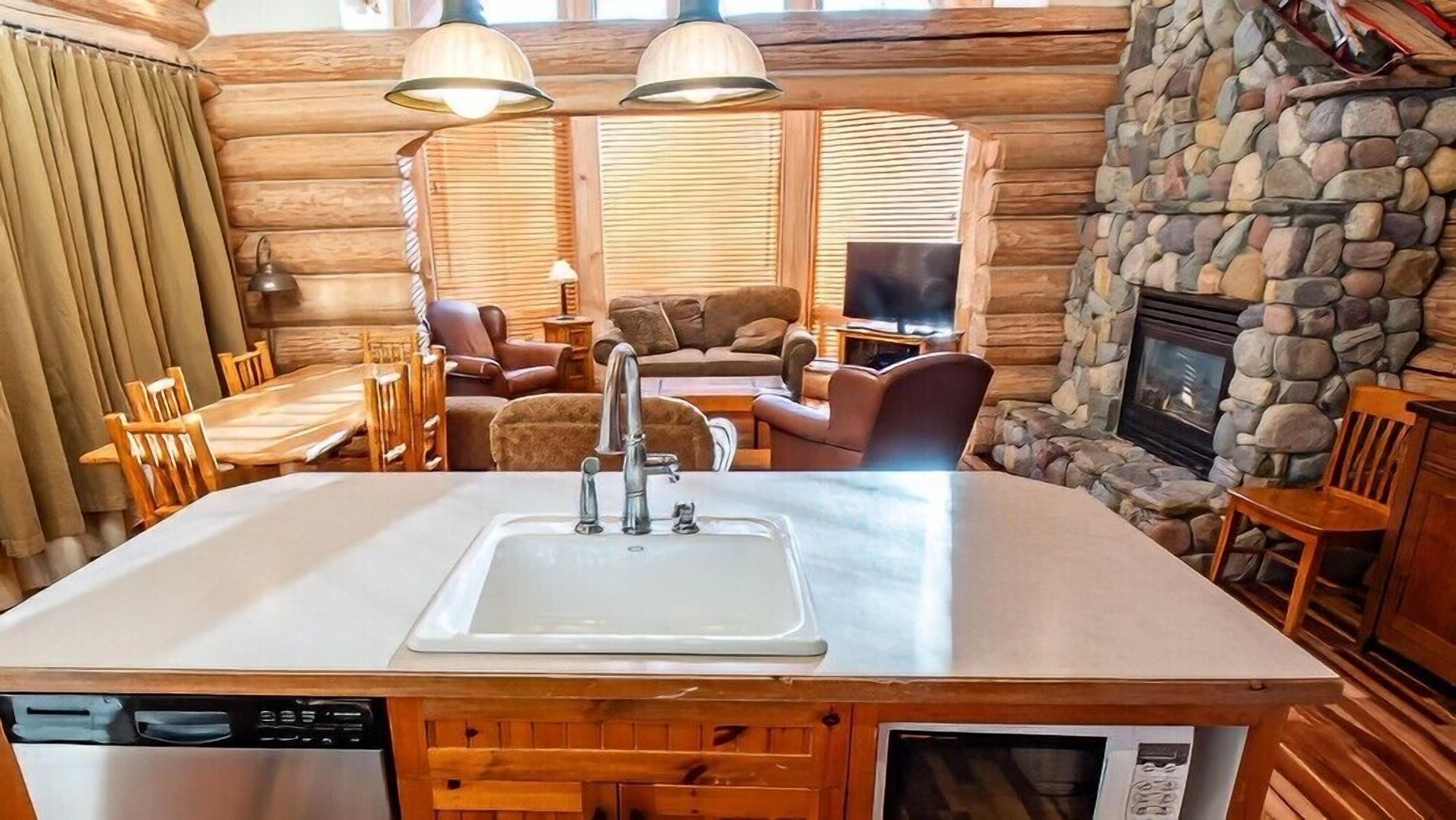 A log cabin interior with a kitchen area featuring a sink, wooden cabinets, and a dishwasher. There is a stone fireplace, a seating area with a couch and chairs, and a television.