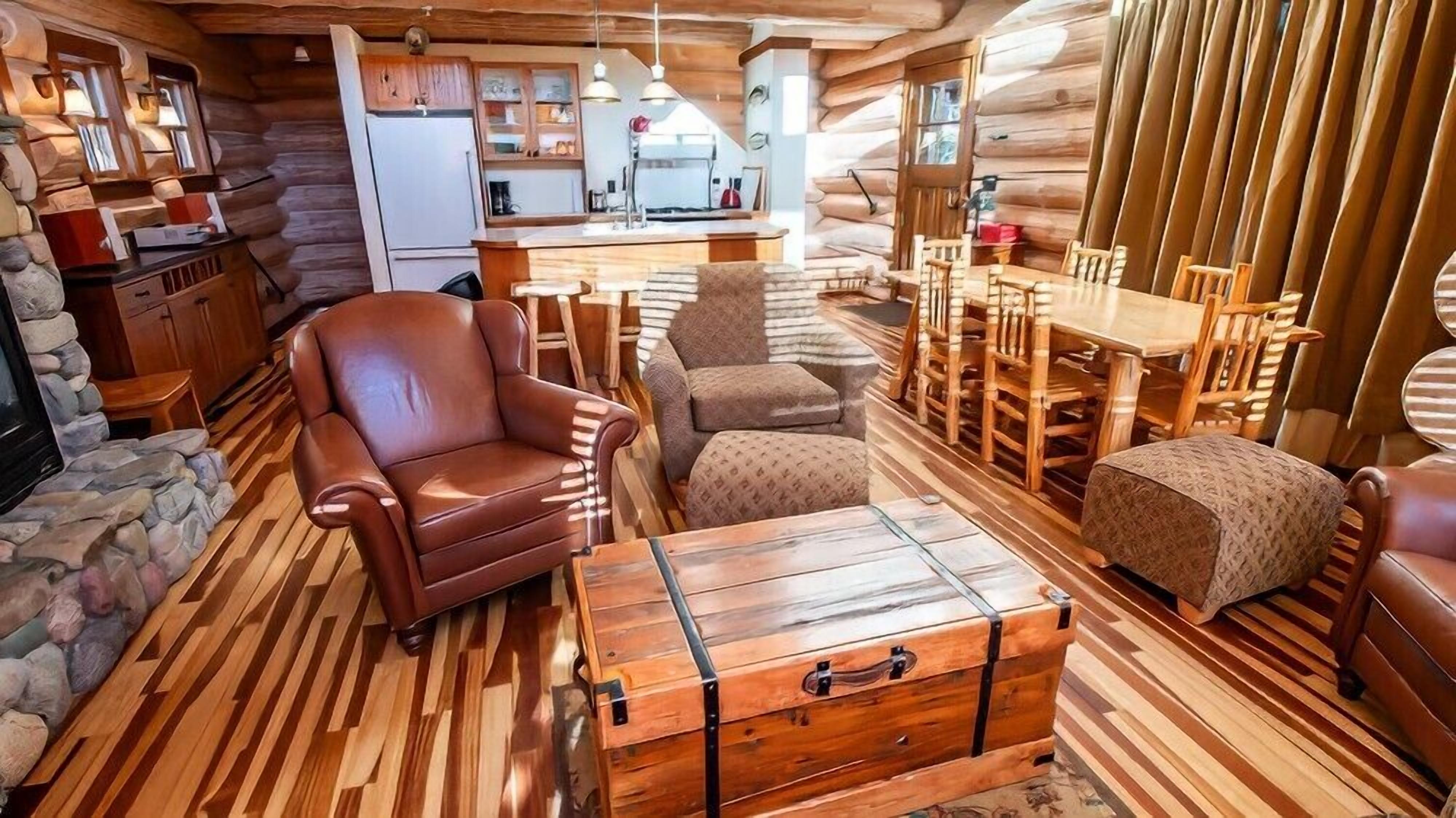 A log cabin interior with a dining area, a kitchen, and a cozy seating area with a leather armchair and ottoman.