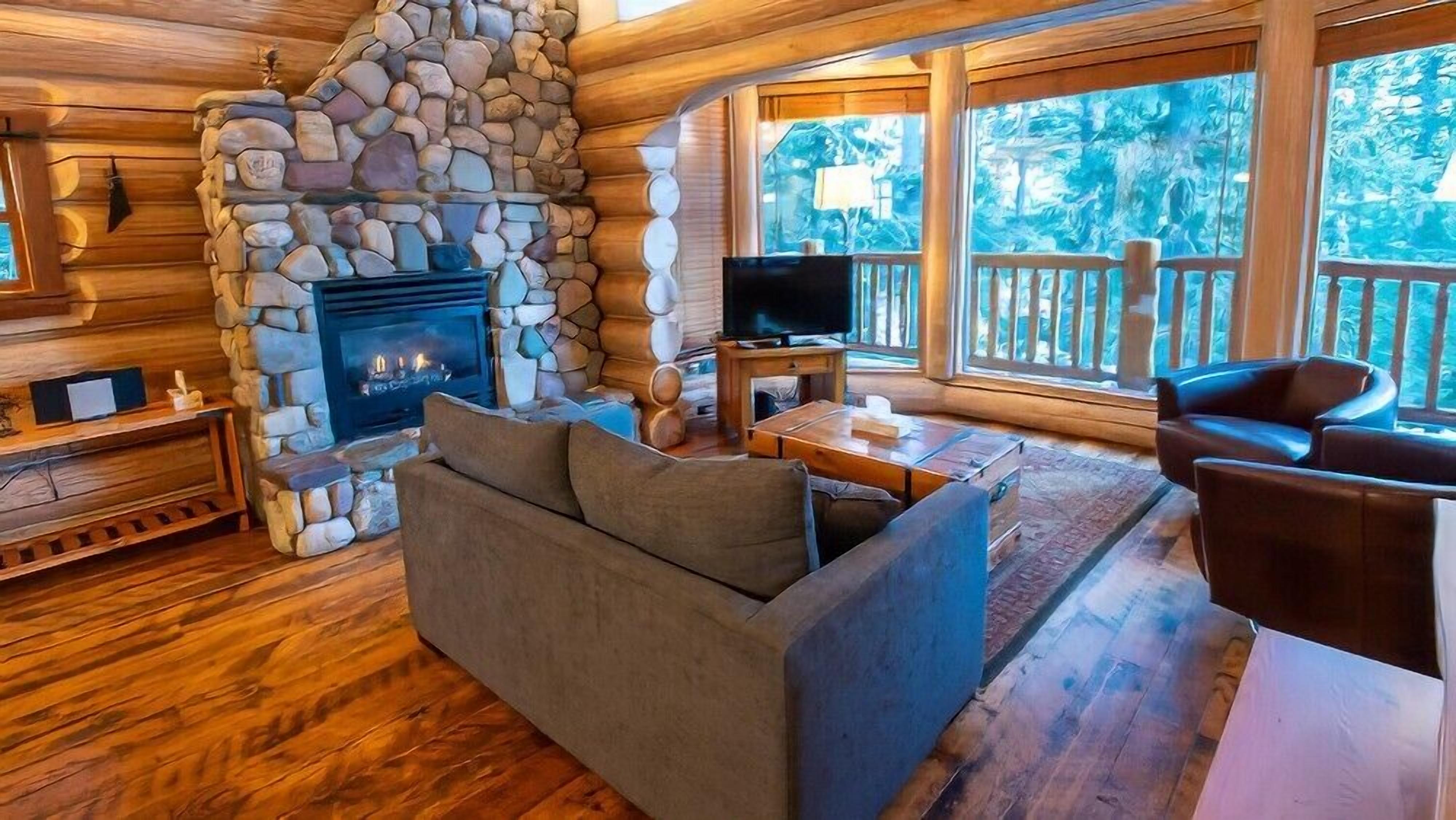 A cozy cabin interior with a stone fireplace, wooden furniture, and large windows offering a view of the forest.