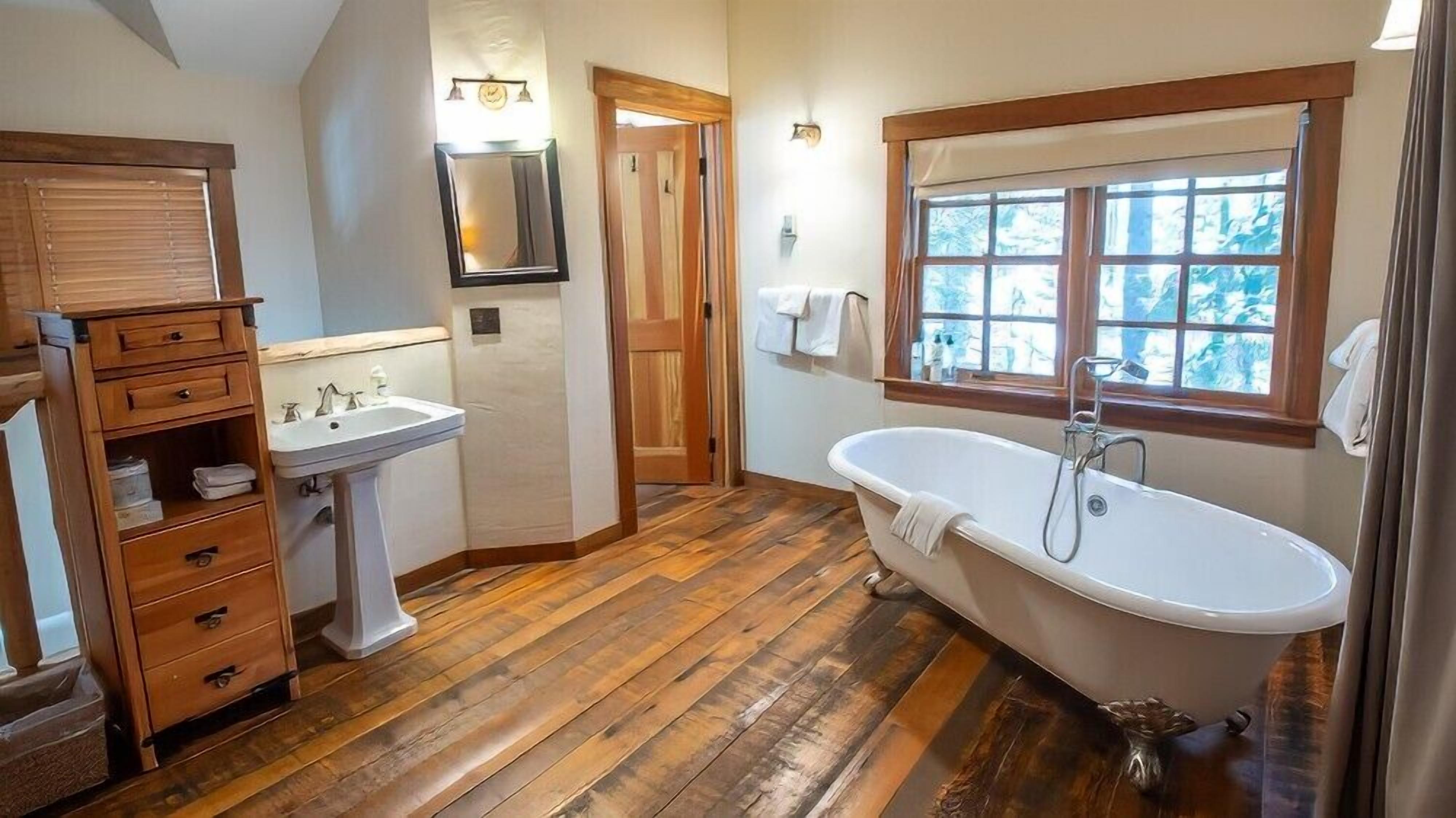 Un baño con suelo de madera, una bañera independiente, un lavabo pedestal y un armario de madera con cajones.