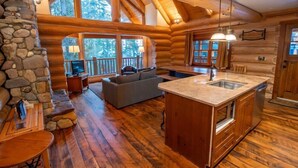 Ein Blockhaushaus-Interieur mit Steinwand, Holzboden, Kamin aus Stein, einer Couch, einem Fernseher und einer Küche mit großer Kochinsel.