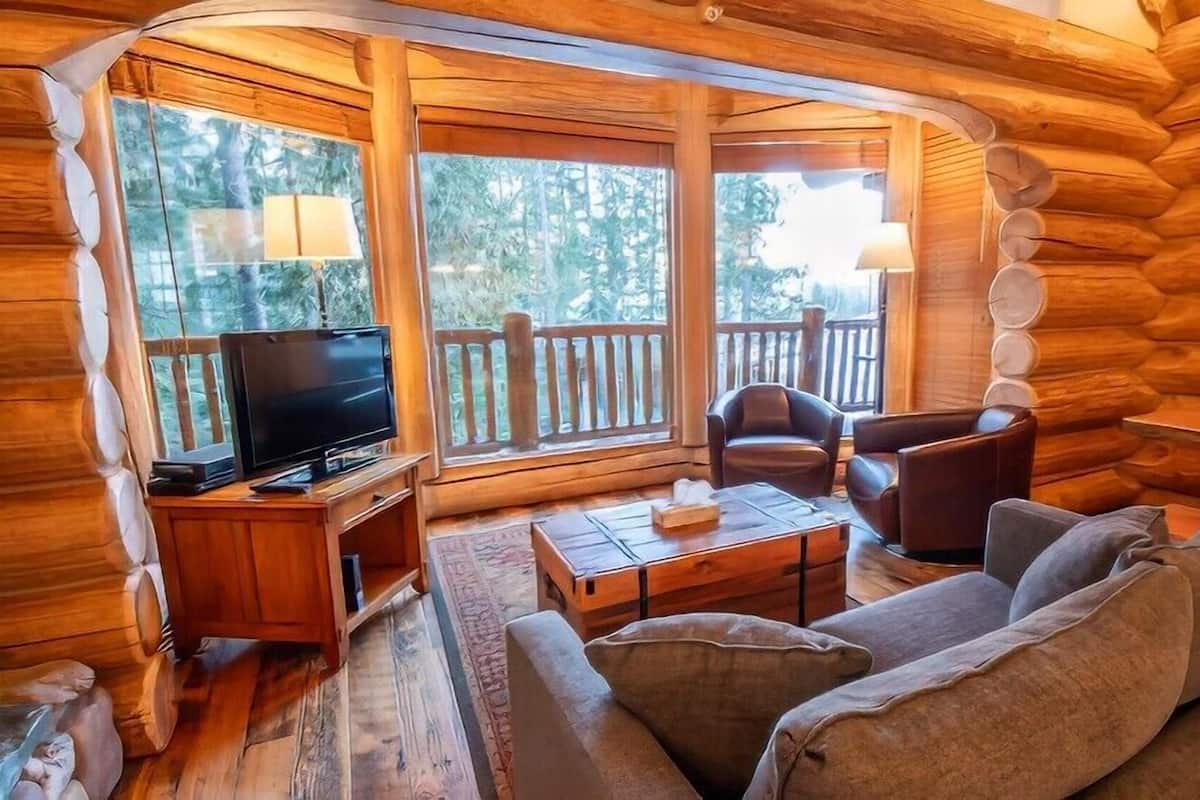 Ein Holzblockhaus-Innenraum mit Holzboden, einem Fernseher, einer Couch, zwei Sesseln und einem großen Fenster mit Blick auf Bäume.