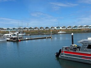Un port de plaisance où sont amarrés plusieurs bateaux, une rangée de maisons de plage en arrière-plan et un ciel bleu dégagé.