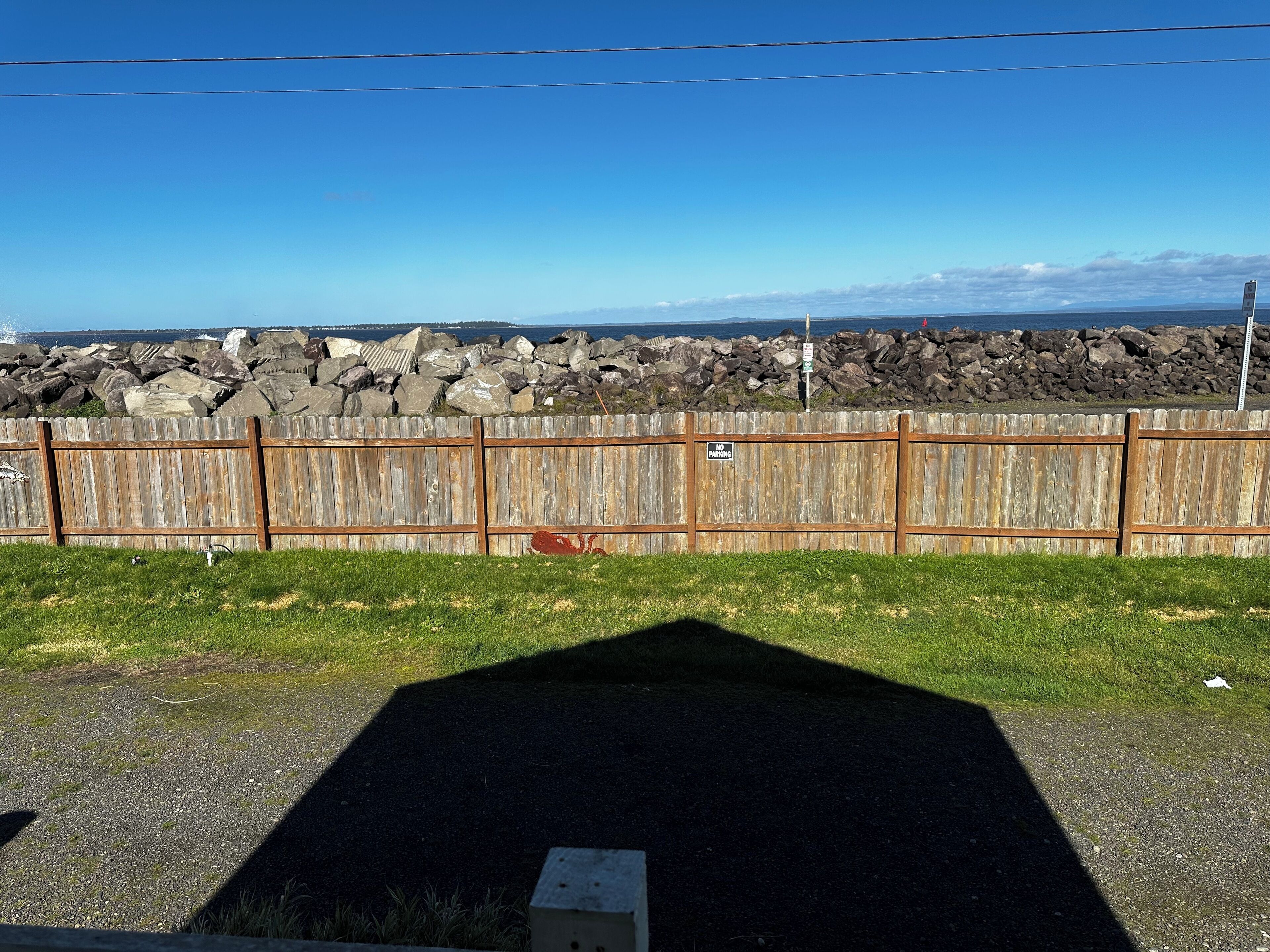 A wooden fence with a 'No Parking' sign, a gravel area, and a view of the ocean and rocks in the background.